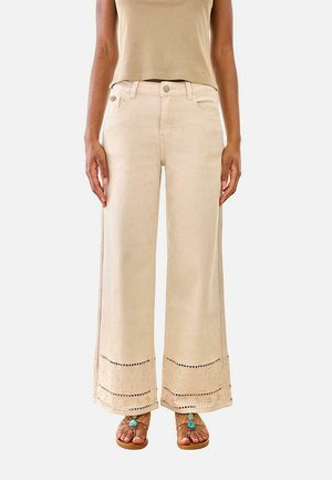 Mujer con pantalones anchos beige con detalle de encaje en los bajos, combinados con top sin mangas beige y sandalias con acentos turquesa.