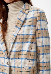Blazer de cuadros marrón y azul con cuello de muesca, cierre de botones y un corte estructurado, superpuesto a un suéter de cuello alto de canalé beige.