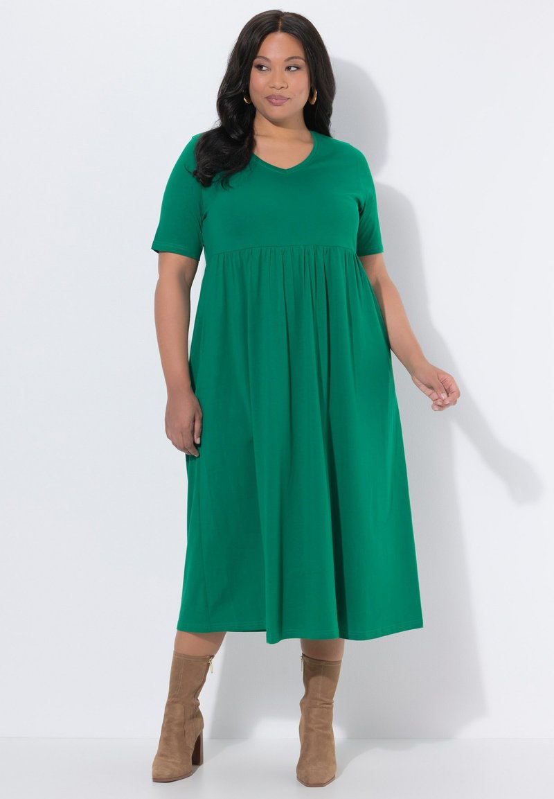 Mujer con un vestido midi verde de manga corta y botines de ante color marrón con tacón, posando frente a un fondo blanco.