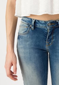 Jeans de mezclilla en azul medio con un acabado desgastado. Presentan cierre de botones, bolsillos frontales y costuras amarillas en contraste.