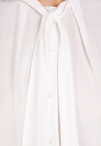 Blouse blanche avec un motif texturé, dotée d'un col noué, d'une fermeture à boutons sur le devant et d'un tissu doux, offrant une apparence légère et aérée.