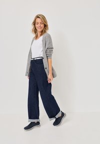 Grauer Strickcardigan über einem weißen Oberteil, kombiniert mit weitgeschnittenen, dunkelblauen Jeans und dunklen Sneakers. Lässiges Outfit mit hochgekrempelten Ärmeln.