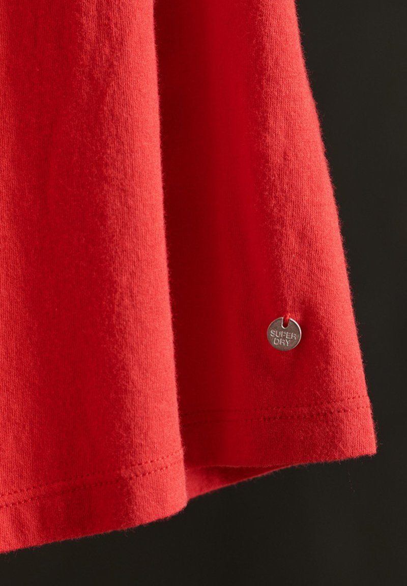 T-shirt in cotone rosso con una texture morbida, caratterizzato da un'etichetta circolare argento con il marchio "SUPER DRY" vicino all'orlo.