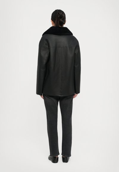 Zwart leren jack met een brede zwarte bontkraag, rechte snit en subtiele textuur. Gecombineerd met donkergrijze broek en platte schoenen.