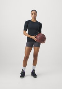 Žena v čiernom športovom tričku a šedých šortkách drží basketbal a stojí na bielom pozadí, pričom má na sebe biele ponožky a čierne tenisky.