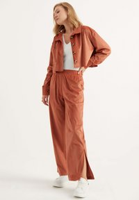 Gemustertes Cord-Set in Rostfarbe bestehend aus einer kurvenförmigen, knopfbelementierten Jacke und lockeren, hochgeschnittenen Hosen. Texturierten Stoff mit seitlichen Schlitzen.