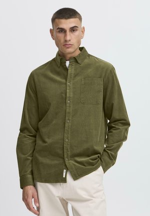 Ung mand iført olivengrøn skjorte med knapper og brystlomme samt lyse beige bukser, stående mod en ensfarvet hvid baggrund.