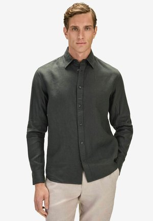 Mand iført en mørkegrøn langærmet skjorte med knapper og beige bukser, stående med den ene hånd i lommen mod en ensfarvet baggrund.