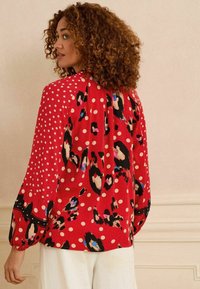 Sarkans blūze ar punktiņiem un leopardu rakstu. Garas, puffa piedurknes ar teksturētām akcentiem pie aprocēm.