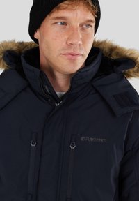 Parka isolée marine avec capuche en fausse fourrure, poches zippées et patch logo. Dotée d'un col montant et d'un tissu durable.