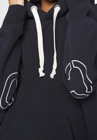 Mörkblå hoodie med framficka, vita dragsnören och kontrasterande vita konturer på ärmarna, med en mjuk textur.