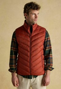 Červená zateplená vesta so zapínaním na zips, s chevronovým prešívaním, stojatým golierom a bočnými vreckami, vrstvená cez kockovanú košeľu.