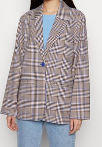 Žena oblečená v hnědo-bílo-modrém kostkovaném blejzru se zeleným knoflíkem, pod ním má světle modré triko a světle modré džíny.