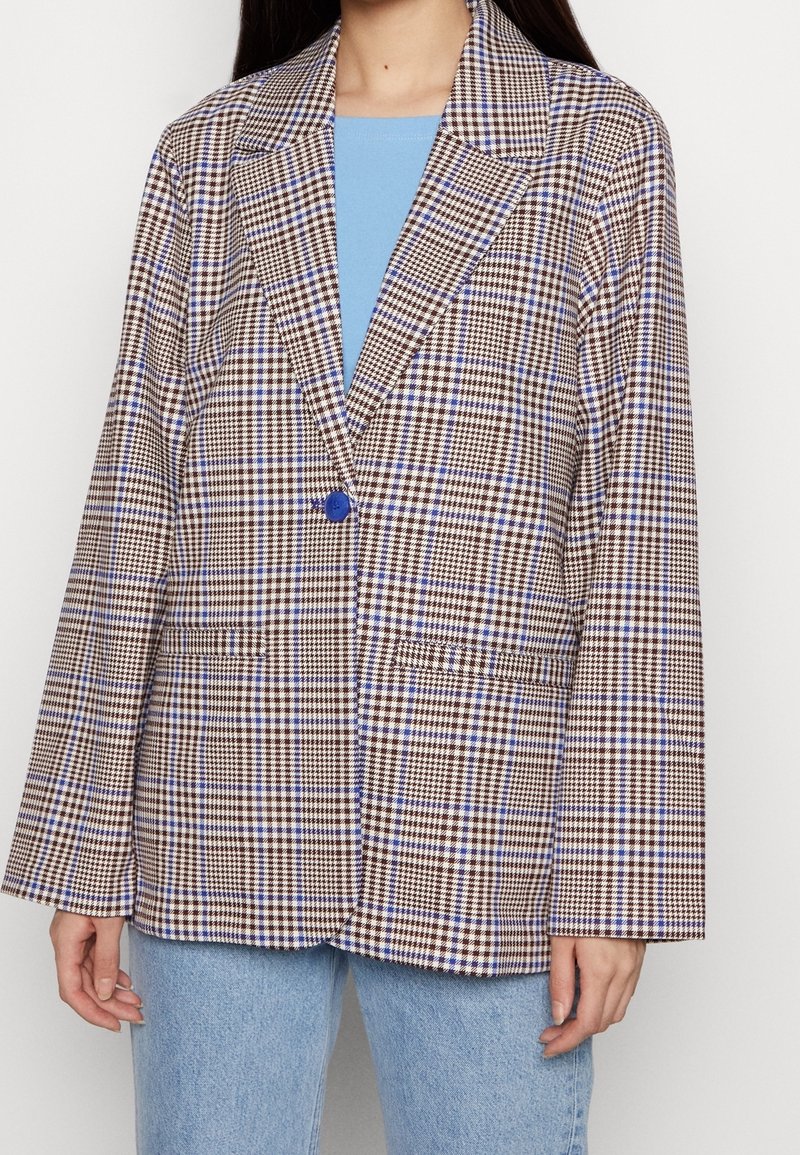 Žena oblečená v hnědo-bílo-modrém kostkovaném blejzru se zeleným knoflíkem, pod ním má světle modré triko a světle modré džíny.