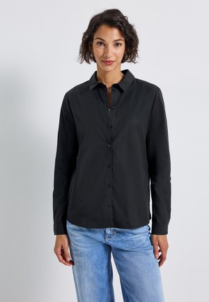 Donna con capelli corti e ricci, indossante una camicia nera con bottoni e jeans azzurri chiari, in piedi contro uno sfondo grigio chiaro uniforme.