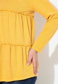 Geel langemouwtop met textuur, gegatherde taille-naad en een ontspannen pasvorm. Gecombineerd met donkerblauwe jeans, hand rustend aan de zijkant.