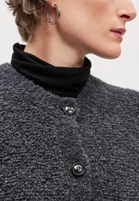 Hallukas halli tekstuuriga jakk kolme musta nupuga, seljas musta kõrge kaelusega särk. Kaasas hõbedane rõngaskõrvarõngas kuldse aktsendiga.