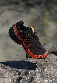 Le scarpe da trail Salomon nere e arancioni presentano una tomaia resistente, un sistema di allacciatura dinamico, una suola robusta e dettagli testurizzati per migliorare la presa.
