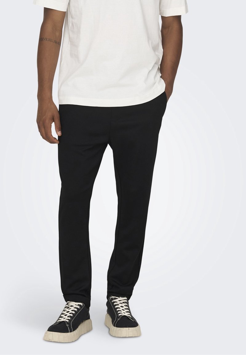 Zwarte tapering broek gemaakt van zachte stof, met een gladde textuur. Gecombineerd met zwarte sneakers en een wit t-shirt voor contrast.
