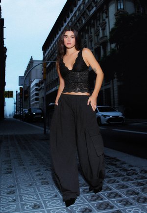 Une femme pose sur un trottoir en ville, vêtue d’un haut noir en dentelle et d’un pantalon à jambes larges. Des bâtiments et une voiture se trouvent derrière elle.