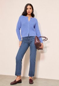 Hele sinine kudumiga kardigan, mille esiküljel on nööbid, koos sirgete jalgevahe teksade ja pruuni mokassiiniga. Hoides burgundi punast kolmnurkset käekotti.