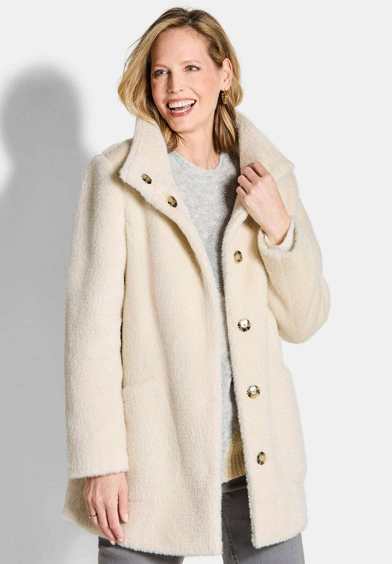 Cremefarbener Wollmantel mit weicher Textur, ausgestattet mit einem Reverskragen und auffälligen Knopfakzenten, präsentiert mit den Händen in den Taschen.