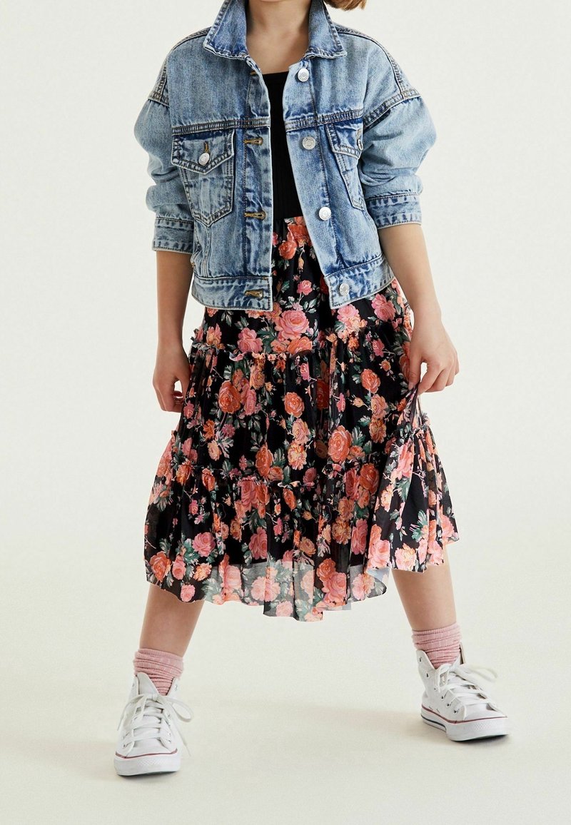 Enfant portant une veste en denim bleu, un haut noir, une jupe midi fleurie, des chaussettes roses et des baskets blanches, debout jambes écartées sur fond blanc.