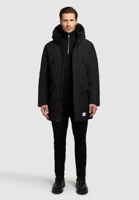 Black insulated parka with a hood, front zip, and snap fasteners. Features two side pockets and subtle logo tag at the hem.