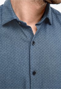 Hombre con una camisa de botones azul de patrones y cuello, mostrando el cuello y la barba de varios días.