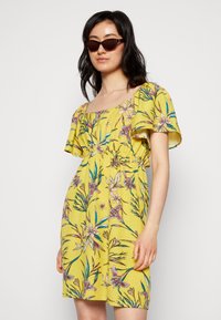 Vestido floral amarelo com mangas curtas e esvoaçantes e um decote quadrado. Apresenta uma cintura franzida com padrões botânicos multicoloridos por toda a peça.
