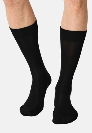 Gambe con calzini neri semplici di lunghezza media fino al polpaccio su uno sfondo grigio chiaro, che mostrano la trama e la vestibilità sui piedi e sulle gambe inferiori.