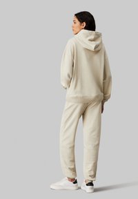 Sudadera beige con una gran capucha, puños y dobladillo acanalados, combinada con pantalones de chándal beige. Llevada con zapatillas blancas con un acento negro.