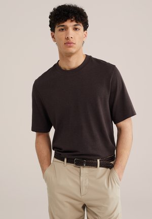 Jeune homme aux cheveux bouclés portant un t-shirt noir à manches courtes, un pantalon beige et une ceinture tressée foncée, debout les mains dans les poches.