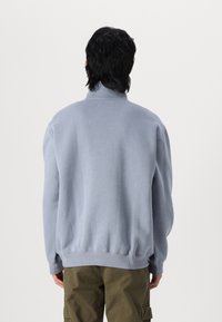 Light blue sweatshirt with a high collar, ribbed cuffs and hem, and a relaxed fit, worn over olive green cargo pants.