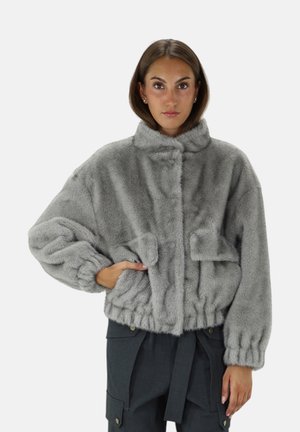 Vrouw draagt een lichtgrijze faux fur jas met elastische manchetten en zoom, staand met één hand in de zak tegen een witte achtergrond.