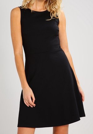 Femme portant une robe noire sans manches, longueur genou, avec un corsage ajusté et une jupe évasée, sur un fond uni.