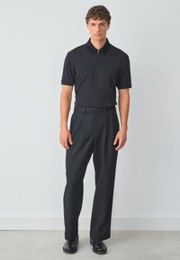 Camisa polo negra con cuello con cremallera y detalles texturizados en negro, combinada con pantalones holgados negros y zapatos negros.