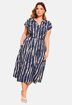 Moteris dėvi tamsiai mėlyną ir baltą midi ilgio su vertikaliomis juostelėmis su trumpomis rankovėmis ir diržu juosmens srityje, pozuoja viena ranka kišenėje, avėdama sandalai.