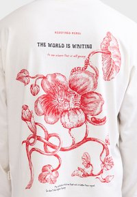 Retro di una camicia bianca con un'illustrazione floreale rossa e testo in nero e rosso: "REDEFINED REBEL. IL MONDO STA ASPETTANDO di vedere dove tutto questo porterà."