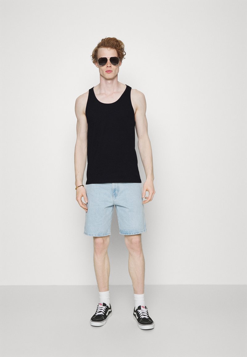 Débardeur noir en coton, shorts en denim bleu clair, baskets noires avec semelles blanches, et lunettes de soleil surdimensionnées. Le modèle se tient contre un fond neutre.