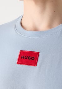 T-shirt bleu clair à col rond avec un patch rectangulaire rouge centré sur la poitrine affichant le texte "HUGO" en lettres noires.