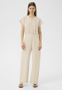 Combinaison en lin beige avec des manches courtes, un d�écolleté en V et une taille élastique. Le design présente des jambes larges et fluides ainsi qu'une texture douce.