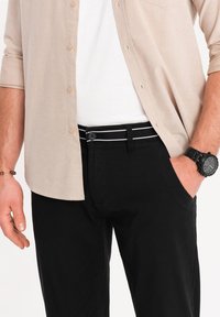 Pantalones negros de corte ajustado, con una cinturilla a rayas. Camisa de botones en un tono beige claro, llevada sobre una camiseta blanca, con un reloj negro.