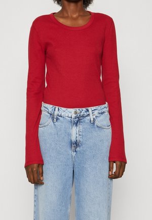 Henkilö pukeutuneena kapeaan punaisen pitkähihaiseen paitaan ja vaalean sinisiin korkeavyötäröisiin farkkuihin, seisomassa yksinkertaisen taustan edessä.