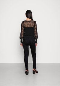 Top negro con mangas de encaje translúcidas y escote con forma de ondas, combinado con jeans ajustados negros y zapatos de tacón alto, visto desde atrás.