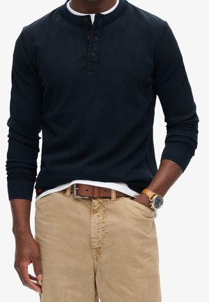 Marineblå Henley-skjorte med lange ærmer, tre-knaps åbning og ribbet tekstur, kombineret med khakifarvede bukser og et brunt bælte. Guldbelagt armbåndsur synligt.