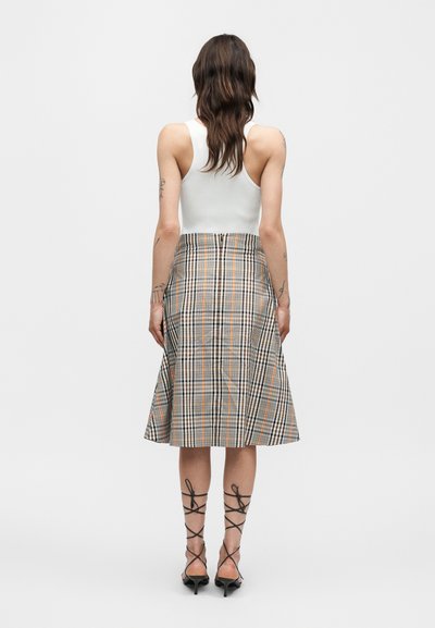 Γυναίκα με μακριά καστανά μαλλιά που φορά λευκό αμάνικο τοπ, καρώ φούστα γραμμή A μεσαίου μήκους και μαύρα πέδιλα με λουράκια, με την πλάτη στραμμένη σε λευκό φόντο.