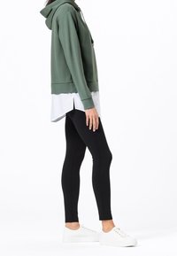 Zielona bluza z kapturem o luźnym kroju, noszona na białej koszuli z bocznymi rozcięciami, zestawiona z czarnymi legginsami i białymi trampkami.