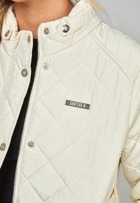 Quilted cream jacket featuring a high collar, silver hardware, and a SIKSILK badge on the chest. Smooth texture with a fitted design.