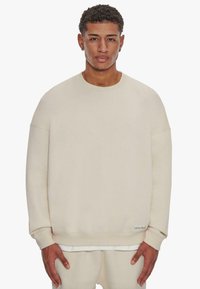 Beige huppari, joka on valmistettu pehmeästä kankaasta. Siinä on pyöreä kaula-aukko, pudotetut olkapäät ja ribbireunukset. Minimalistinen muotoilu, jossa on tarratiedot.